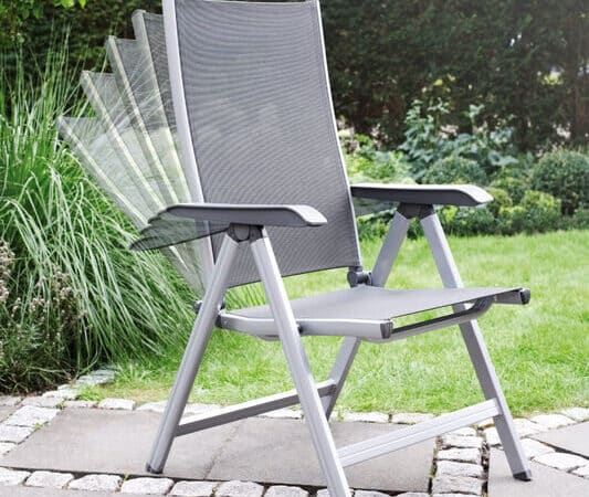 Grey outdoor garden chair with an adjustable reclining back on a stone patio, aluminum frame and mesh seat, with a green hedge in the background.