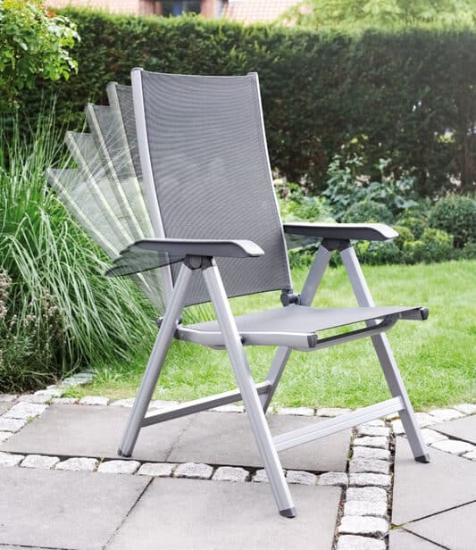 Grey outdoor garden chair with an adjustable reclining back on a stone patio, aluminum frame and mesh seat, with a green hedge in the background.