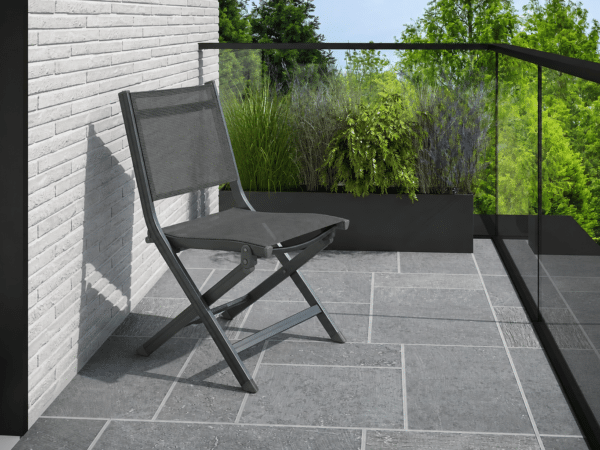 Dark gray folding mesh chair on a modern tiled balcony with a brick wall and glass railing, greenery in the planter beyond.