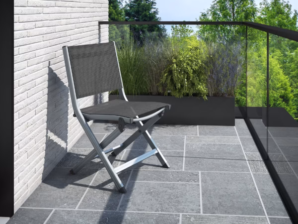 Metal lounge chair on a tiled balcony beside a white brick wall, with a glass railing and green trees beyond.