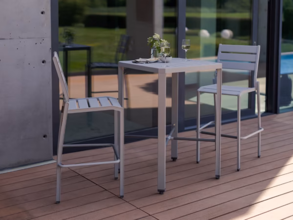 Outdoor dining setup on a wooden deck: a white metal table with two matching chairs, set with wine glasses, a small flower arrangement, and bread near glass doors by a pool.