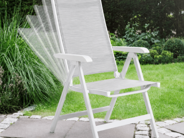 White folding outdoor chair on a stone patio in a green garden setting.