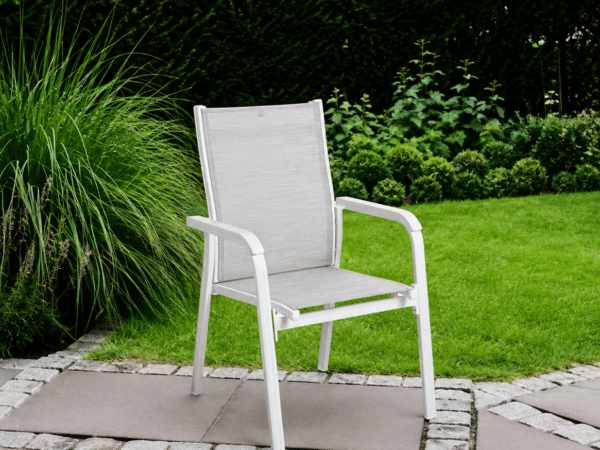 White outdoor patio chair with grey fabric seat on a stone-tiled patio beside lush green plants.